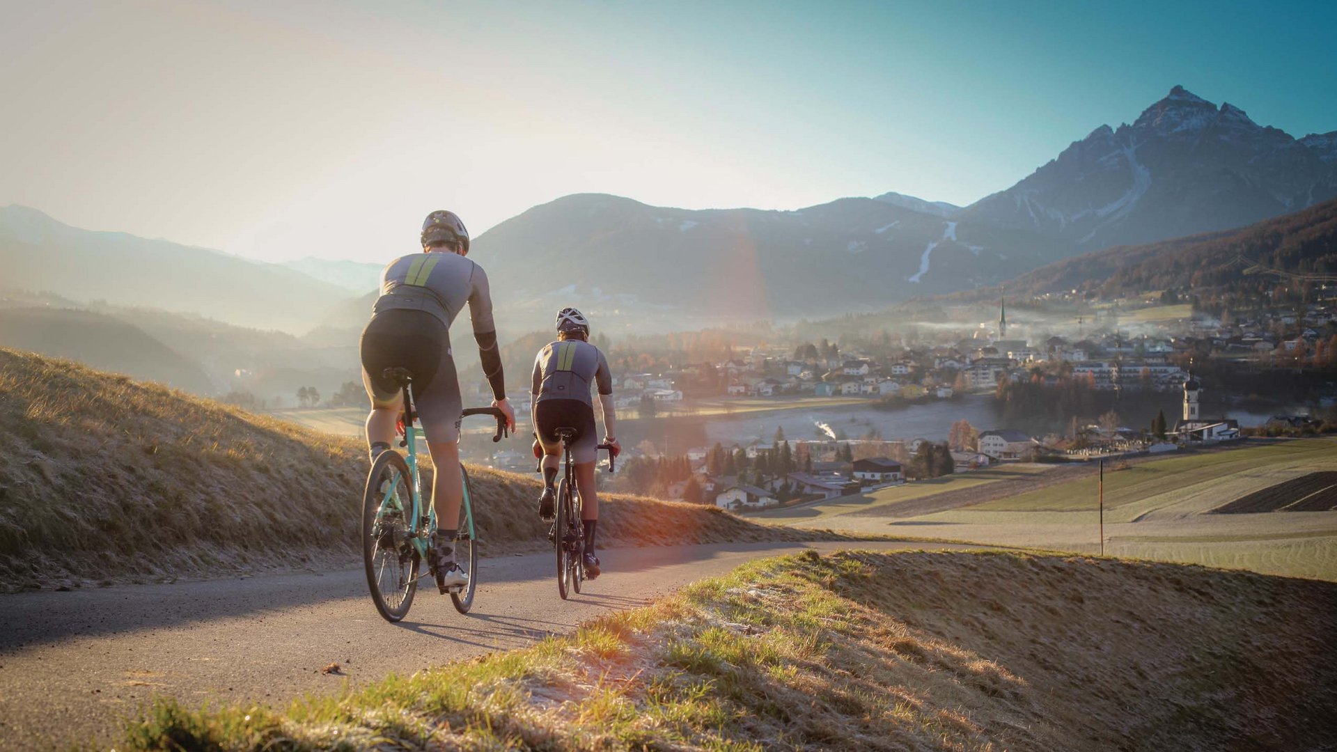 Mobilità e MTB nello Stubai Due ciclisti su strada di campagna con montagne e paese sullo sfondo all'alba