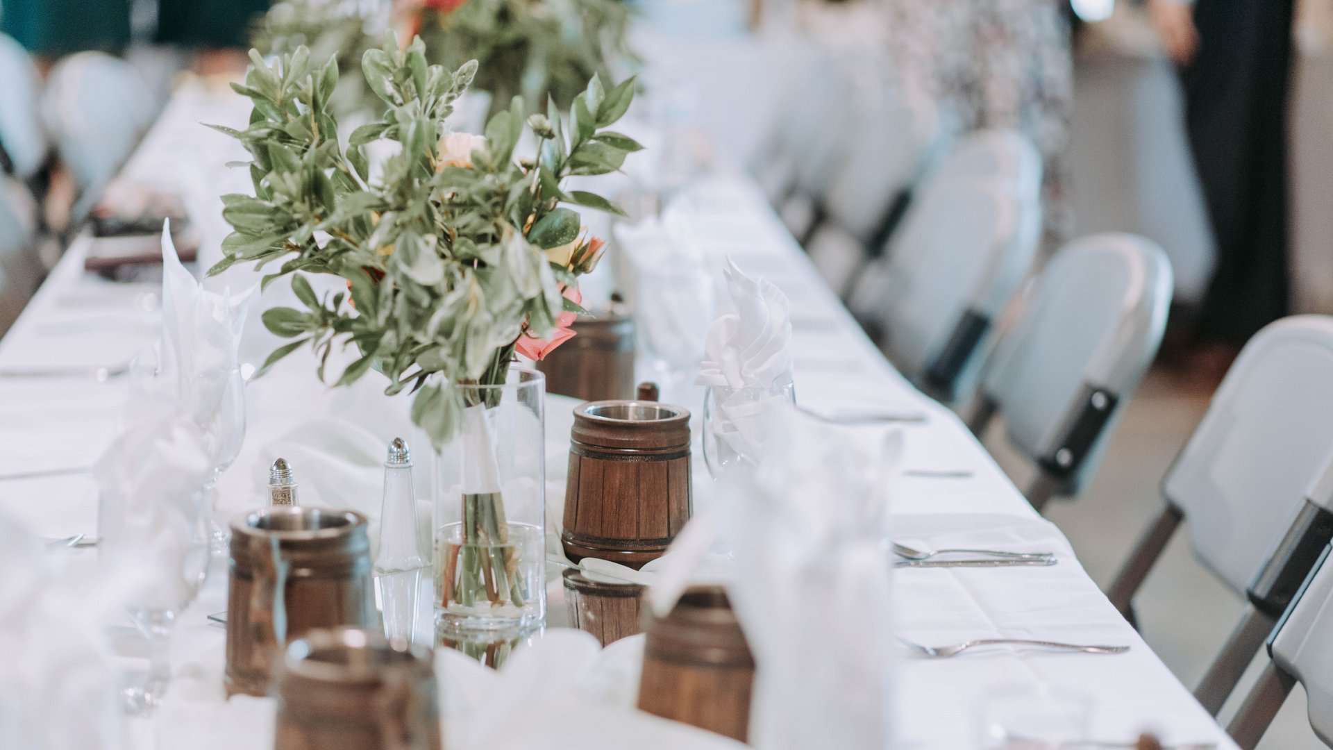 Tavolo lungo con tovaglia bianca, vaso di fiori e bicchieri di legno per evento