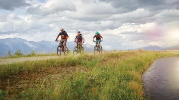 News (Teaser) Drei Mountainbiker fahren auf einem Weg neben einem Gewässer in den Bergen