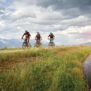 News (Teaser) Drei Mountainbiker fahren auf einem Weg neben einem Gewässer in den Bergen