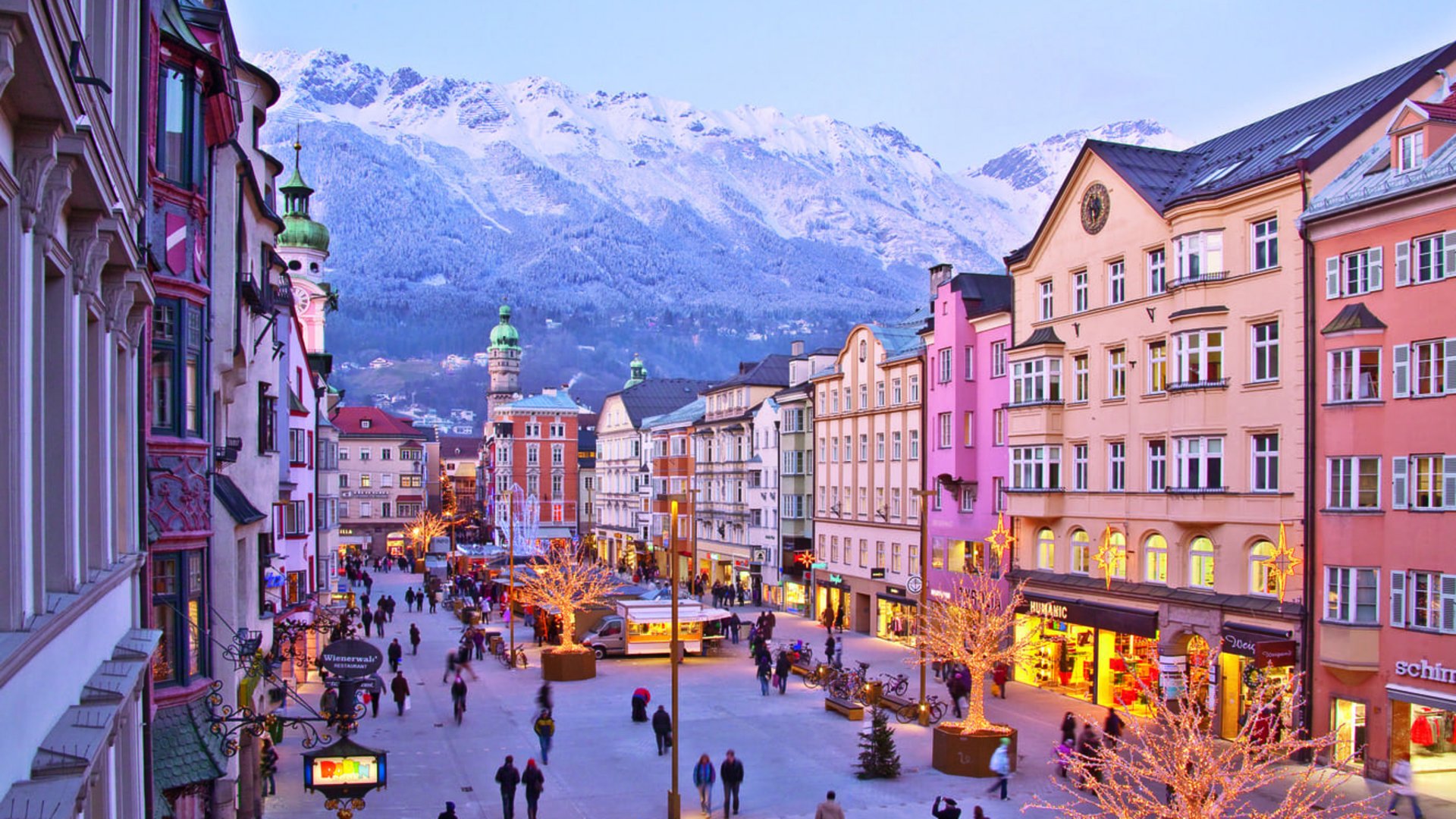 Belebte Fußgängerzone mit beleuchteten Bäumen und schneebedeckten Bergen im Hintergrund