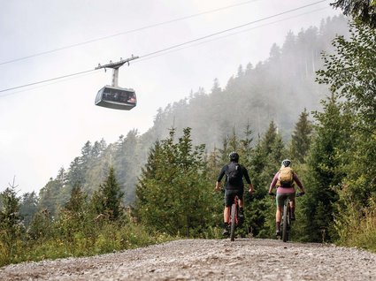 Parkhotel Matrei: Mountainbikehotel in Tirol Zwei Mountainbiker auf Waldweg mit Seilbahn im Nebel über ihnen