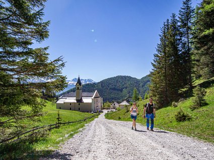 Wanderhotel in Tirol: herrliche Berge und Täler Zwei Wanderer auf einem Waldweg nahe einer Kapelle in den Bergen bei klarem Himmel