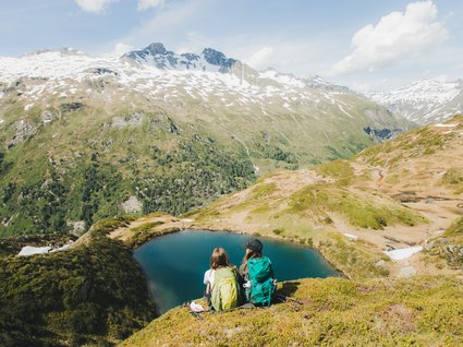 Wanderhotel in Tirol: herrliche Berge und Täler Zwei Wanderer mit Rucksäcken sitzen am Bergsee vor schneebedeckten Gipfeln