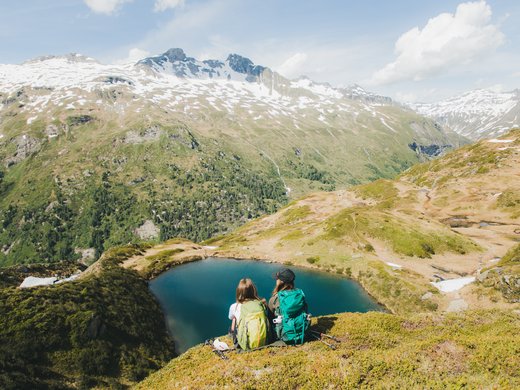 Sporthotel in Tirol: Aktivurlaub im Parkhotel Matrei Zwei Wanderer mit Rucksäcken sitzen am Bergsee vor schneebedeckten Gipfeln