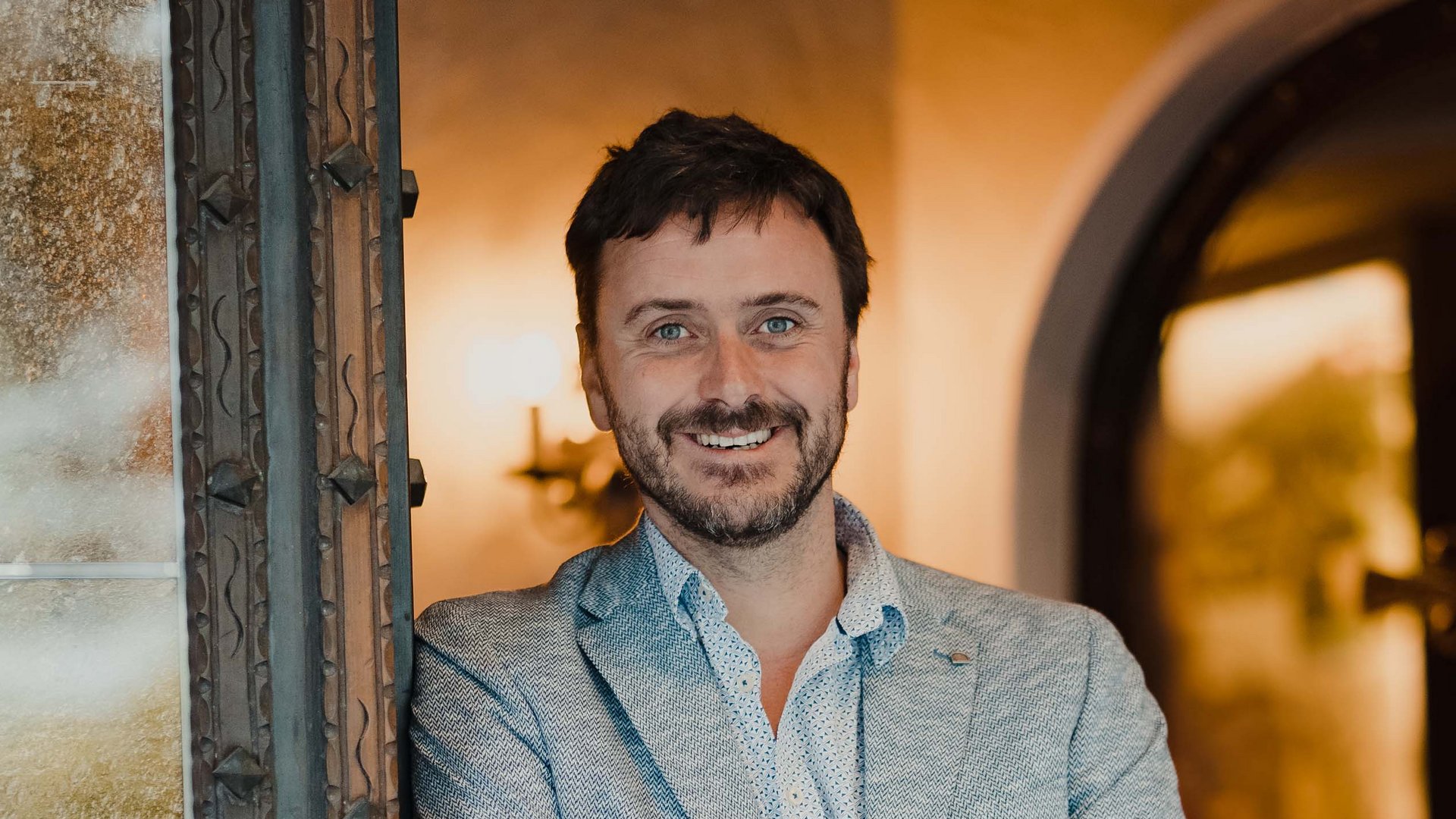 Smiling man leaning against door frame with arms crossed in warmly lit room