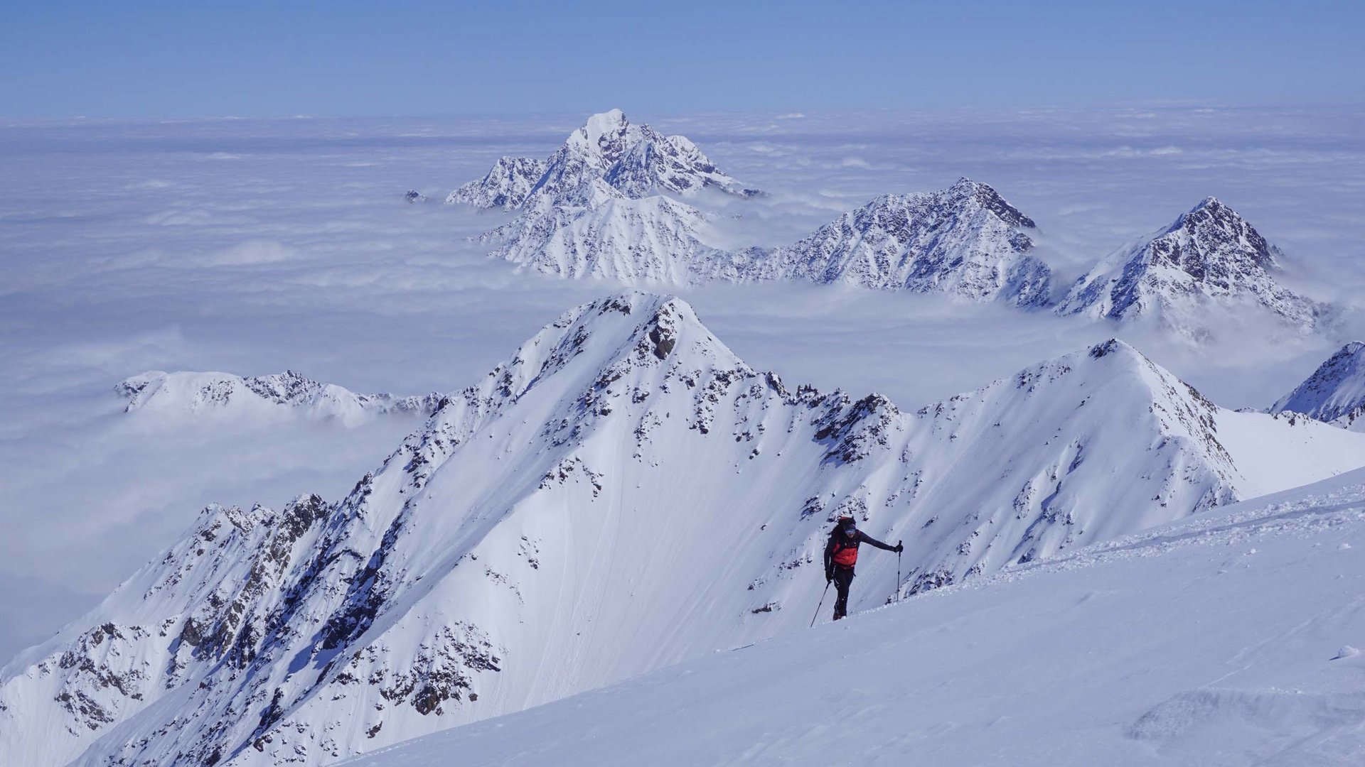 Bergsteiger im roten Rucksack wandert durch verschneite Alpen mit Wolkenschicht darunter