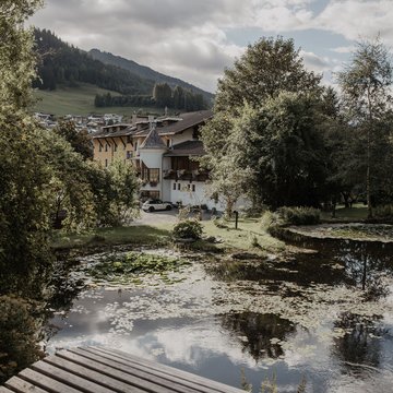 Neuigkeiten aus dem Motorradhotel in Tirol Teich mit Seerosen vor Häusern und bewaldeten Hügeln bei bewölktem Himmel