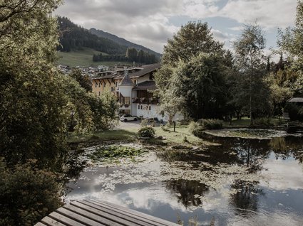 Wanderhotel in Tirol: herrliche Berge und Täler Teich mit Seerosen vor Häusern und bewaldeten Hügeln bei bewölktem Himmel