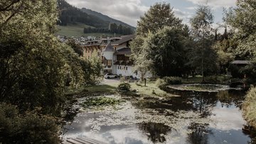 News (Liste) Teich mit Seerosen vor Häusern und bewaldeten Hügeln bei bewölktem Himmel