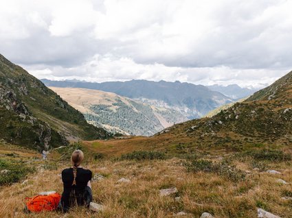 Wanderhotel in Tirol: herrliche Berge und Täler Frau sitzt auf Wiese und blickt auf bewaldete Berge und bewölkten Himmel