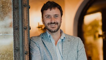 News (Liste) Mann mit Haaren und Bart lächelt, trägt hellgrauen Anzug und steht an Türrahmen