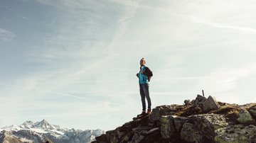 News (Liste) Mann mit Rucksack steht auf felsigem Berggipfel mit schneebedeckten Bergen im Hintergrund