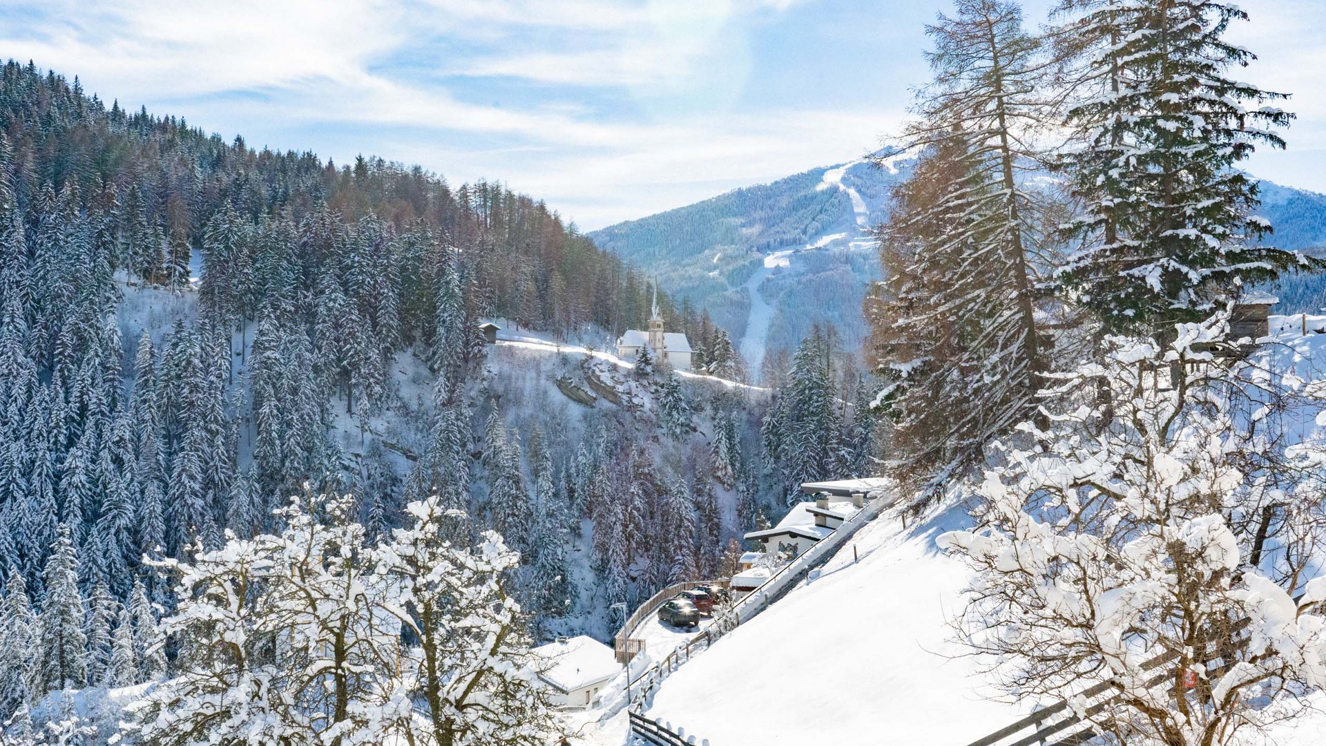 Snowy winter landscape with forest, mountains, and houses on a sunny day