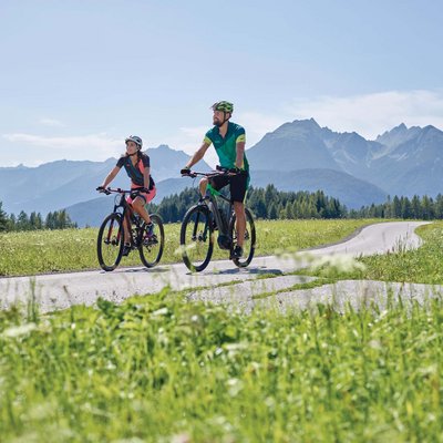 News (Liste) Zwei Radfahrer fahren auf Landstraße mit Bergen und Wiesen im Hintergrund