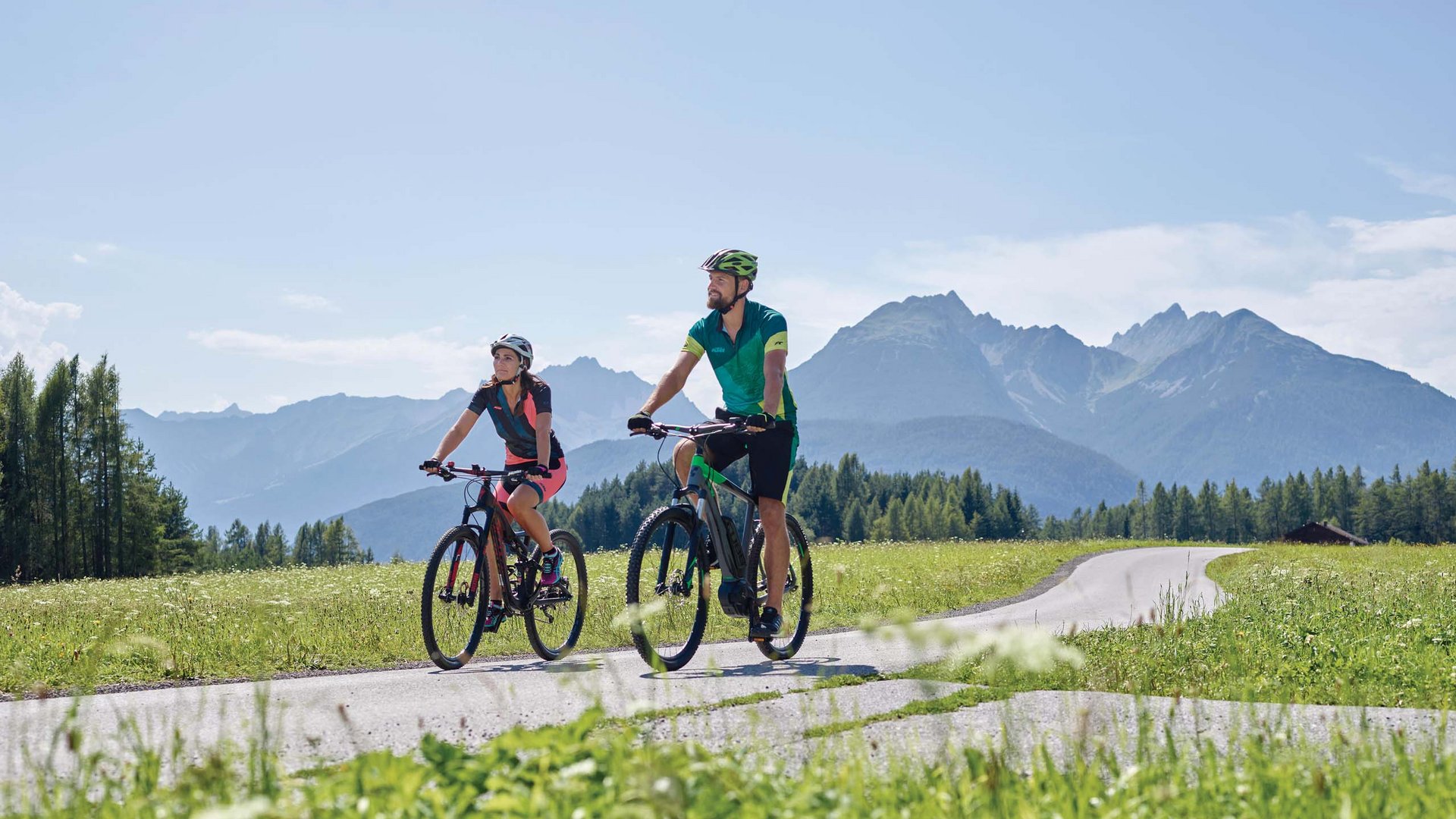 Parkhotel Matrei: Mountainbikehotel in Tirol Zwei Radfahrer fahren auf Landstraße mit Bergen und Wiesen im Hintergrund