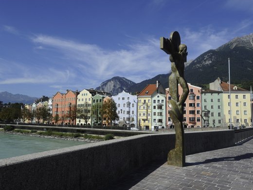 Ausflugstipps aus unserem Hotel: Innsbruck und Umgebung Bronzestatue an Flussufer mit bunten Häusern und Bergen im Hintergrund