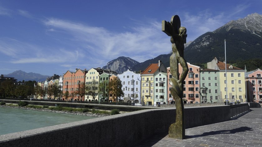 Zeitreise durch die Epochen Bronzestatue an Flussufer mit bunten Häusern und Bergen im Hintergrund